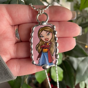 May include: A silver necklace with a rectangular pendant depicting a cartoon woman. The pendant has a pink background and a silver beaded edge. The woman has brown hair, a red jacket, blue jeans, and is wearing a silver necklace.