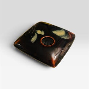 May include: A square ceramic flower frog with a glossy dark brown glaze and subtle cream-colored mottled accents. The center features a circular opening containing a black metal pin holder with numerous upright needles for arranging stems.