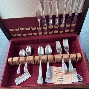 May include: A vintage silverware set in a wooden case with a red velvet lining. The set includes spoons, forks, and knives with ornate handles. The case contains a label that reads "1847 Rogers Bros. American's Finest Silverplate".