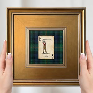 Könnte beinhalten: Ein gerahmtes Kunstwerk mit einem Spielkarten-Design. Die Karte, ein Kreuz-Ass, zeigt einen Golfspieler beim Schwung. Das Kunstwerk ist vor einem grün-blauen Schottenmuster-Hintergrund platziert und in einem goldfarbenen Rahmen gerahmt.