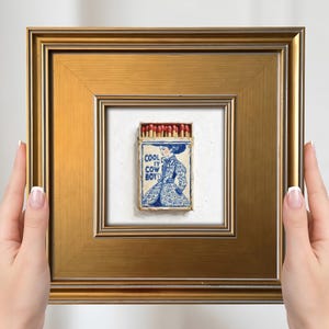 Puede incluir: Una obra de arte enmarcada que presenta una caja de cerillas con una ilustración azul y blanca de una persona con un vestido, con el texto "COOL COW BOY". La caja de cerillas está colocada sobre un fondo blanco y enmarcada en un marco dorado.