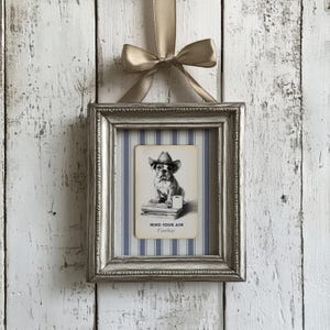 May include: A framed print featuring a bulldog wearing a cowboy hat, with the text "MIND YOUR AIM Cowboy". The print is set against a blue and white striped background, within a silver frame, and is suspended by a gold ribbon.