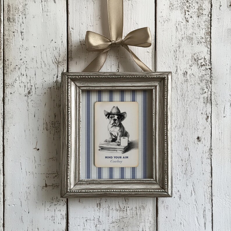 May include: A framed print featuring a bulldog wearing a cowboy hat, with the text "MIND YOUR AIM Cowboy". The print is set against a blue and white striped background, within a silver frame, and is suspended by a gold ribbon.
