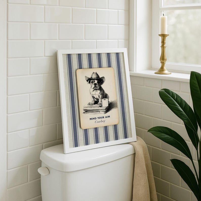 May include: A framed print featuring a bulldog wearing a cowboy hat, with the text "MIND YOUR AIM Cowboy". The print is set against a blue and white striped background and a white frame. The print is displayed on a white toilet.