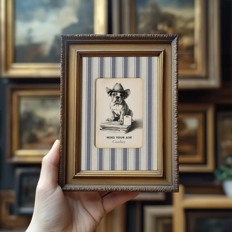 May include: Framed artwork featuring a bulldog wearing a cowboy hat. The artwork is set against a blue and white striped background and framed in a gold-toned frame. The text "MIND YOUR AIM Cowboy" is printed below the image.