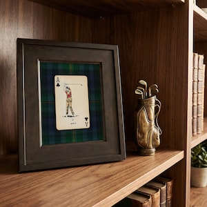 May include: A framed Ace of Clubs playing card with a golfer illustration, set against a green and blue plaid background. A bronze golf bag with clubs sits on a wooden shelf, alongside books and a small potted plant.