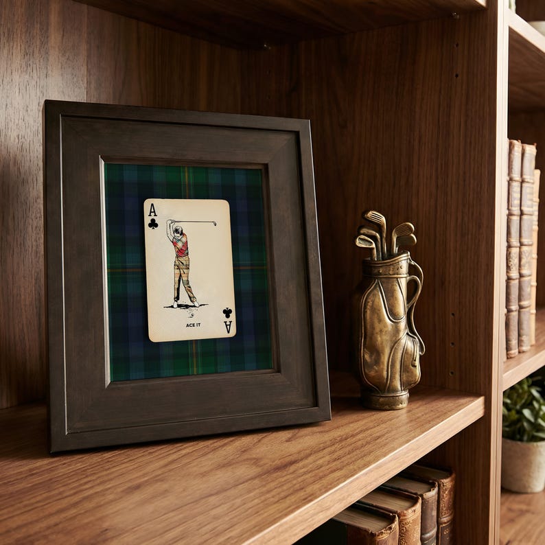 May include: A framed Ace of Clubs playing card with a golfer illustration, set against a green and blue plaid background. A bronze golf bag with clubs sits on a wooden shelf, alongside books and a small potted plant.