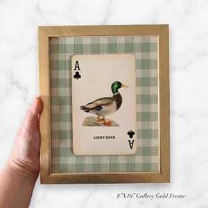 May include: A framed wall art piece featuring an 8 inch by 10 inch gold frame containing a playing card with an illustration of a mallard duck. The card displays the text Lucky Duck and the letter A with a club symbol in the corners.
