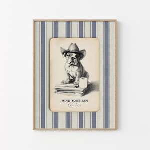 May include: Framed print featuring a bulldog wearing a cowboy hat, sitting on books with a roll of toilet paper. The text "MIND YOUR AIM Cowboy" is below the image. The frame has a striped blue and white pattern.