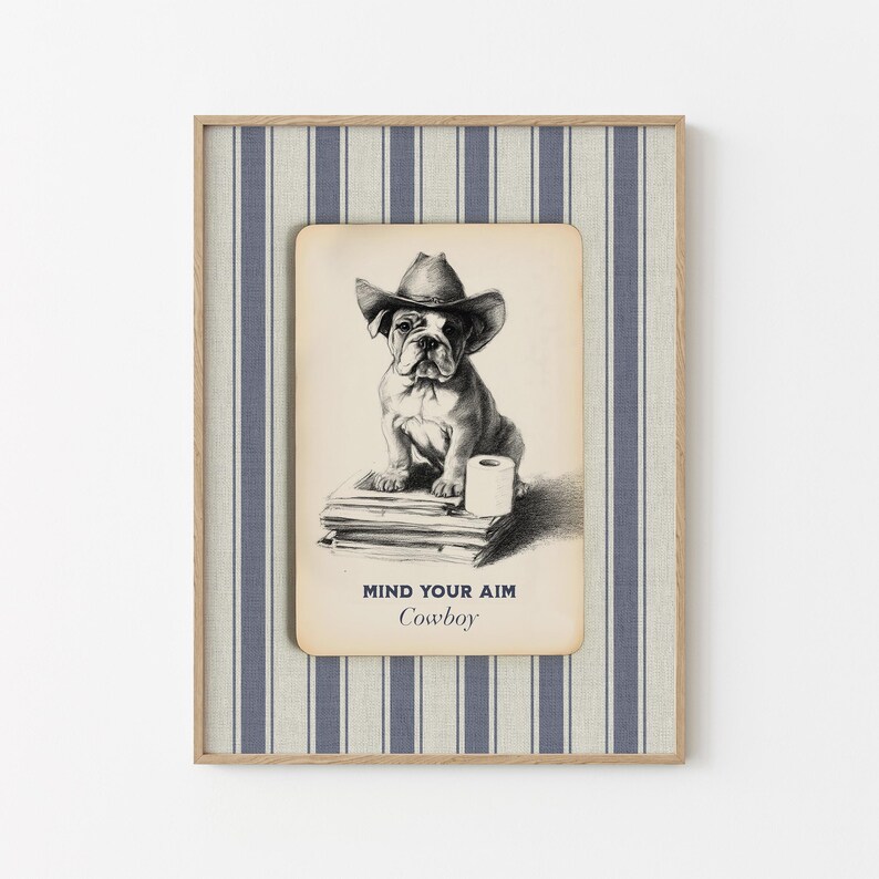 May include: Framed print featuring a bulldog wearing a cowboy hat, sitting on books with a roll of toilet paper. The text "MIND YOUR AIM Cowboy" is below the image. The frame has a striped blue and white pattern.