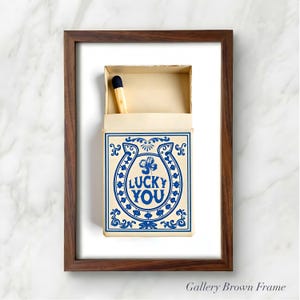 May include: A framed artwork featuring a matchbox with a blue and cream design. The matchbox has a horseshoe and clover graphic with the words "LUCKY YOU". A single match is visible inside the open box. The frame is brown.