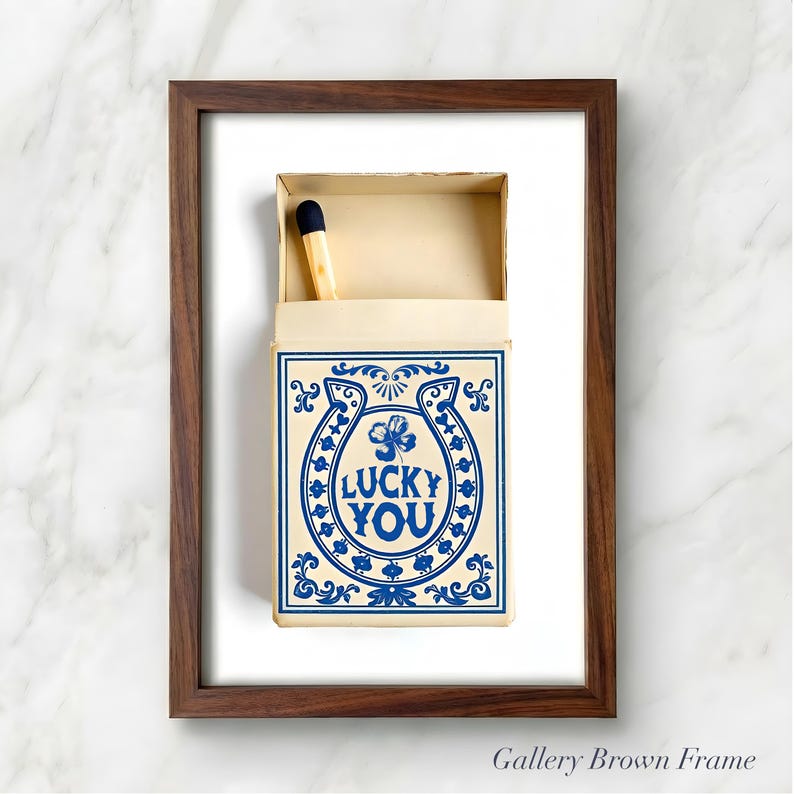 May include: A framed artwork featuring a matchbox with a blue and cream design. The matchbox has a horseshoe and clover graphic with the words "LUCKY YOU". A single match is visible inside the open box. The frame is brown.