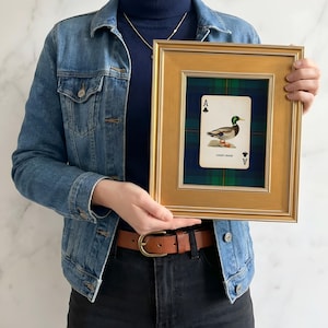May include: A framed playing card featuring an illustration of a duck, labeled "Lucky Duck." The card is set against a green and blue plaid background within a gold-colored frame. The frame is rectangular.