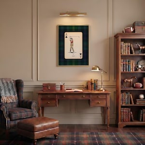 May include: A framed Ace of Clubs playing card featuring a golfer, set against a green and blue plaid background. The room includes a wooden desk, a bookshelf, a plaid armchair, and a leather ottoman, creating a classic, cozy aesthetic.