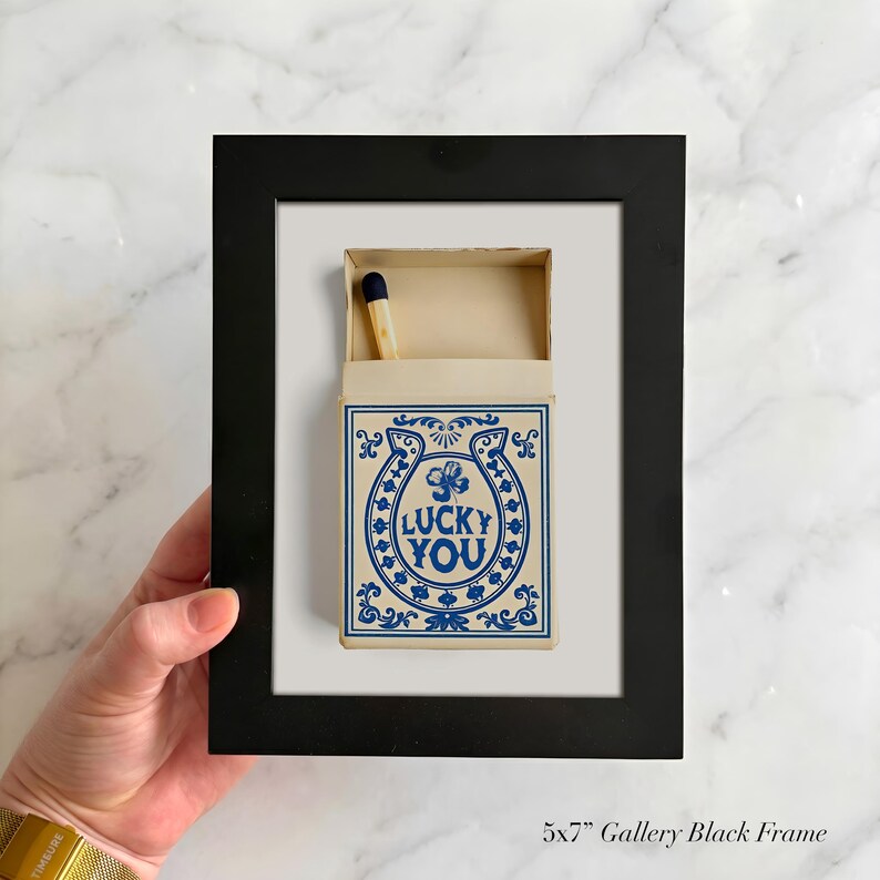 May include: A framed matchbox art piece. The matchbox is cream-colored with a blue horseshoe design and the words "LUCKY YOU." A single match with a blue tip is visible. The frame is black.