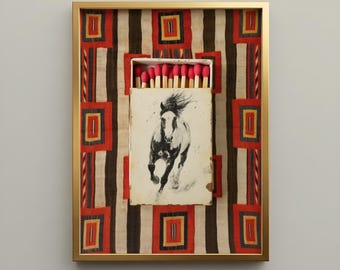 May include: A framed wall art piece featuring a matchbox with a black and white illustration of a running horse. The matchbox contains wooden matches with bright pink tips. The background is a fabric with a geometric pattern of red, black, and yellow.