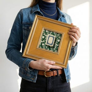 May include: A square, gold-framed artwork featuring a small, rectangular tile with a cream-colored center and a blue border. The tile is set against a green floral patterned fabric. The frame is held by a person wearing a denim jacket.
