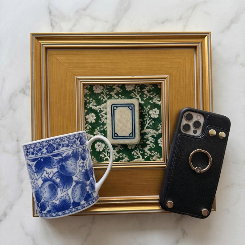 May include: A blue and white ceramic mug with a strawberry design sits next to a black phone case with a ring holder. A framed tile with a blue border is displayed on a green floral background.