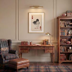 Puede incluir: Una l&aacute;mina de arte enmarcada con un dise&ntilde;o de carta de As de Corazones, una ilustraci&oacute;n de un colimbo y el texto REBEL LOON. La obra se exhibe sobre un escritorio de madera con cajones, una l&aacute;mpara, libros y un sill&oacute;n con una manta a cuadros.