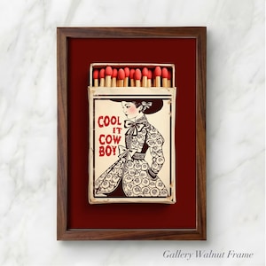 Op de afbeelding: Een ingelijst kunstwerk met een lucifersdoosontwerp. De lucifersdoos toont de tekst "COOL IT COW BOY" en een illustratie van een vrouw in een patroonjurk en hoed. De lijst is van walnoot, tegen een dieprode achtergrond.