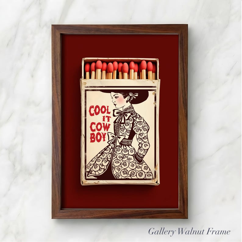 Op de afbeelding: Een ingelijst kunstwerk met een lucifersdoosontwerp. De lucifersdoos toont de tekst "COOL IT COW BOY" en een illustratie van een vrouw in een patroonjurk en hoed. De lijst is van walnoot, tegen een dieprode achtergrond.