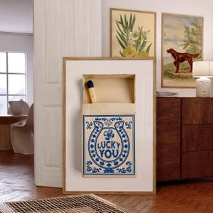 May include: A framed artwork featuring a large matchbox with a single match. The matchbox is cream-colored with a blue horseshoe design and the words "LUCKY YOU". Two framed botanical prints and a dog painting are visible in the background.