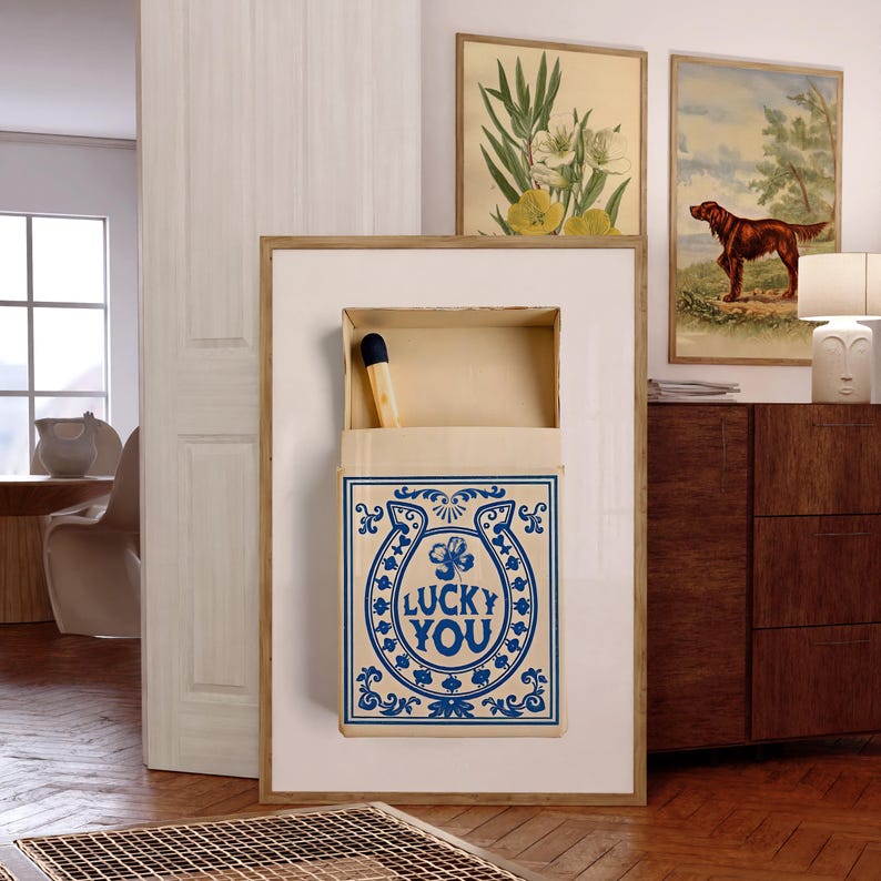 May include: A framed artwork featuring a large matchbox with a single match. The matchbox is cream-colored with a blue horseshoe design and the words "LUCKY YOU". Two framed botanical prints and a dog painting are visible in the background.