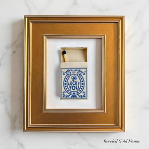 May include: A framed matchbox with a gold beveled frame. The matchbox has a blue and white design with the words "LUCKY YOU" inside a horseshoe. A single match with a black tip is visible.