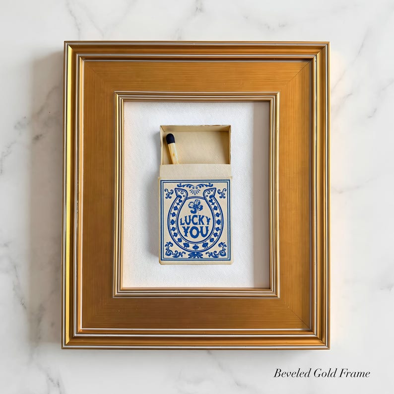 May include: A framed matchbox with a gold beveled frame. The matchbox has a blue and white design with the words "LUCKY YOU" inside a horseshoe. A single match with a black tip is visible.