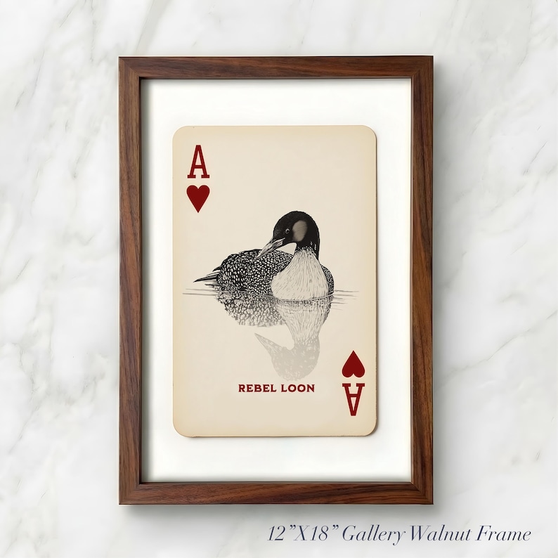 Puede incluir: Una impresi&oacute;n enmarcada de 30,48 cm x 45,72 cm con un dise&ntilde;o de carta de juego que muestra una ilustraci&oacute;n en blanco y negro de un colimbo en el agua. La carta tiene fondo crema, corazones rojos, la letra A en las esquinas y el texto REBEL LOON.