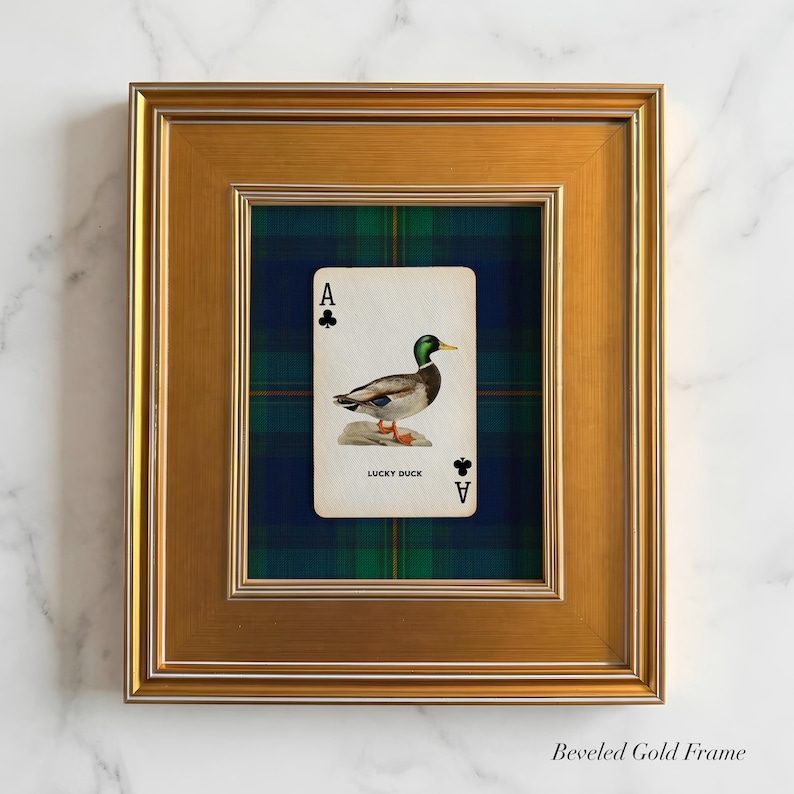 May include: A framed playing card featuring a duck illustration, labeled "Lucky Duck," set against a plaid background. The card is framed in a gold beveled frame, creating a decorative wall art piece.