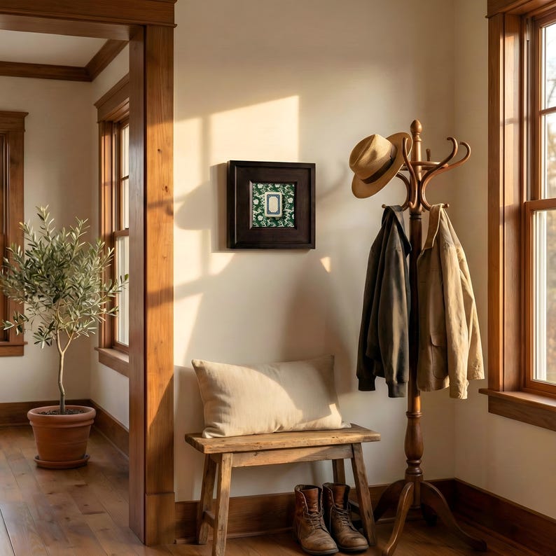 A warm, inviting interior scene featuring a wooden coat rack with two jackets and a hat. An espresso framed print of a white dragon Mahjong tile hangs on the wall, above a wooden bench with a pillow. A potted olive tree and boots complete the cozy entryway.
