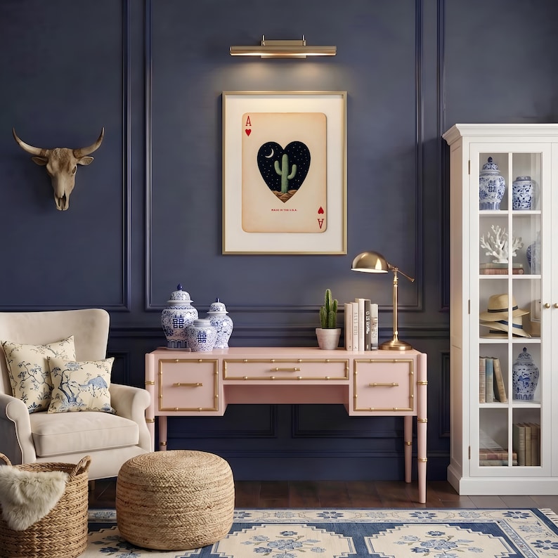 May include: A room with a navy blue wall, a pink desk with gold accents, and a framed playing card art piece. The art features a heart-shaped design with a cactus and a night sky. Decorative items include blue and white vases, a bull skull, and a white cabinet.