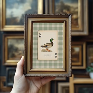 May include: A framed print featuring a playing card design with a duck illustration. The card is the Ace of Clubs, with the words "LUCKY DUCK" printed below the duck image. The frame is made of dark, ornate wood.