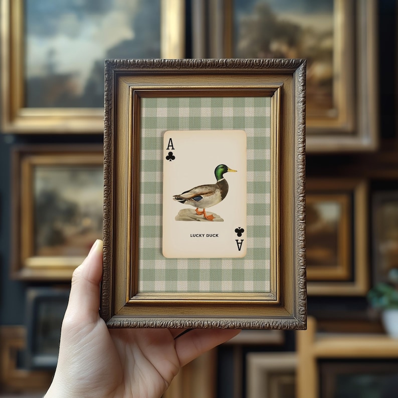May include: A framed print featuring a playing card design with a duck illustration. The card is the Ace of Clubs, with the words "LUCKY DUCK" printed below the duck image. The frame is made of dark, ornate wood.