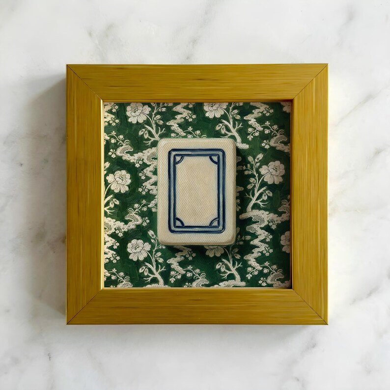 May include: A framed rectangular tile with a cream-colored center and a blue border. The tile is set against a green floral patterned background within a gold-colored wooden frame. The frame is on a white marble surface.