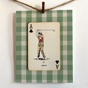 K&ouml;nnte beinhalten: Eine rechteckige Karte mit einer Illustration im Vintage-Stil eines Golfers, der einen Schl&auml;ger schwingt, vor einem gr&uuml;n-wei&szlig;en Vichy-Karo-Hintergrund. Die Karte zeigt den Text ACE IT sowie den Buchstaben A mit einem Kreuz-Symbol.