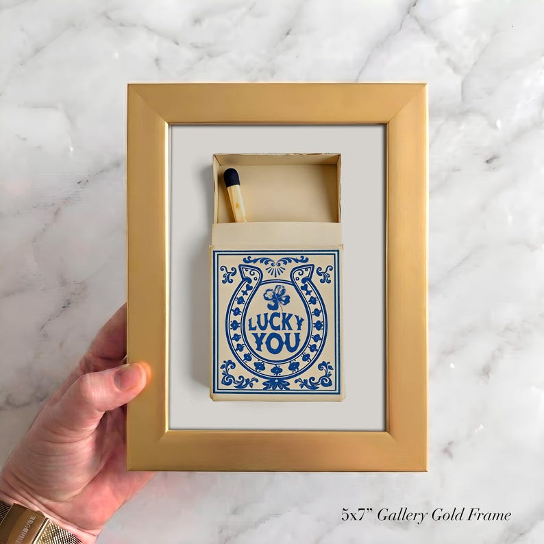 May include: A gold framed print featuring a matchbox with a blue horseshoe design and the words "LUCKY YOU". The matchbox is open, revealing a single match. The frame is labeled "5x7" Gallery Gold Frame".
