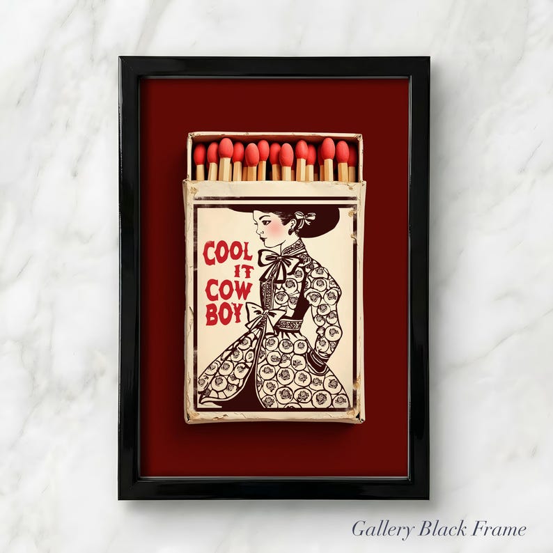 Op de afbeelding: Ingelijst kunstwerk met een lucifersdoos met een vintage illustratie van een vrouw in cowboykleding. Op de lucifersdoos staat "COOL IT COWBOY" in rode tekst. De lucifers hebben rode koppen. Het kunstwerk zit in een zwart frame.