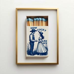 Puede incluir: Una impresión enmarcada de una caja de cerillas con una ilustración azul de una persona con sombrero de vaquero y otra con sombrero de sol tomadas de la mano. El texto HOW LUCKY ARE WE está impreso en azul. La caja contiene cerillas de punta azul.