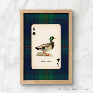 May include: A framed print featuring a playing card with an illustration of a duck. The card is the Ace of Clubs, with the text "LUCKY DUCK" below the duck image. The frame is light wood, set against a green and blue plaid background.