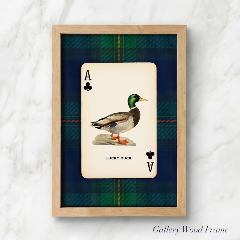 May include: A framed print featuring a playing card with an illustration of a duck. The card is the Ace of Clubs, with the text "LUCKY DUCK" below the duck image. The frame is light wood, set against a green and blue plaid background.