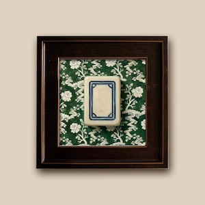 A framed piece featuring a rectangular White Dragon Mahjong tile with a cream-colored center and a blue border. The tile is set against a green floral chinoiserie patterned background and enclosed in a dark brown wooden frame. The artwork is displayed on a neutral-colored wall. Print is 5x5" Slimwood Black-Gold frame is .85" Finished size is 6.5"x6.5"