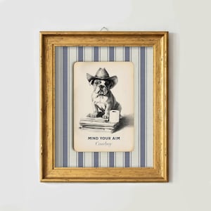 May include: Framed artwork featuring a vintage-style illustration of a bulldog wearing a cowboy hat. The dog sits atop books with a roll of toilet paper. The text "MIND YOUR AIM Cowboy" is below the image. The frame is gold.