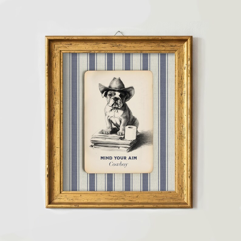 May include: Framed artwork featuring a vintage-style illustration of a bulldog wearing a cowboy hat. The dog sits atop books with a roll of toilet paper. The text "MIND YOUR AIM Cowboy" is below the image. The frame is gold.