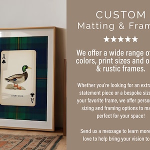 May include: Framed artwork featuring a playing card with a duck illustration, set against a blue and green plaid background. The frame is a light wood color. The text "CUSTOM Matting & Frames" is at the top.