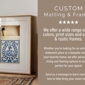 May include: Framed artwork featuring a matchbox design with a blue horseshoe and the text "LUCKY YOU." The artwork is set in a white mat and a light wood frame. The image also includes the text "CUSTOM Matting & Frames."