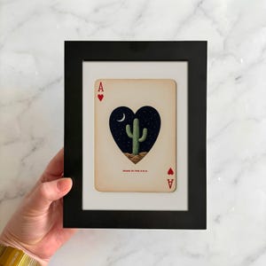 May include: Framed playing card art featuring a cactus in a heart-shaped design under a night sky. The card is framed in a black frame with a white mat. The card reads "MADE IN THE U.S.A." and has the Ace of Hearts symbol.