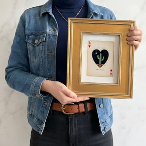 May include: A framed playing card featuring a heart-shaped design with a cactus, moon, and stars. The card is an Ace of Hearts, set in a gold-colored frame. The text "MADE IN THE U.S.A." is visible on the card.