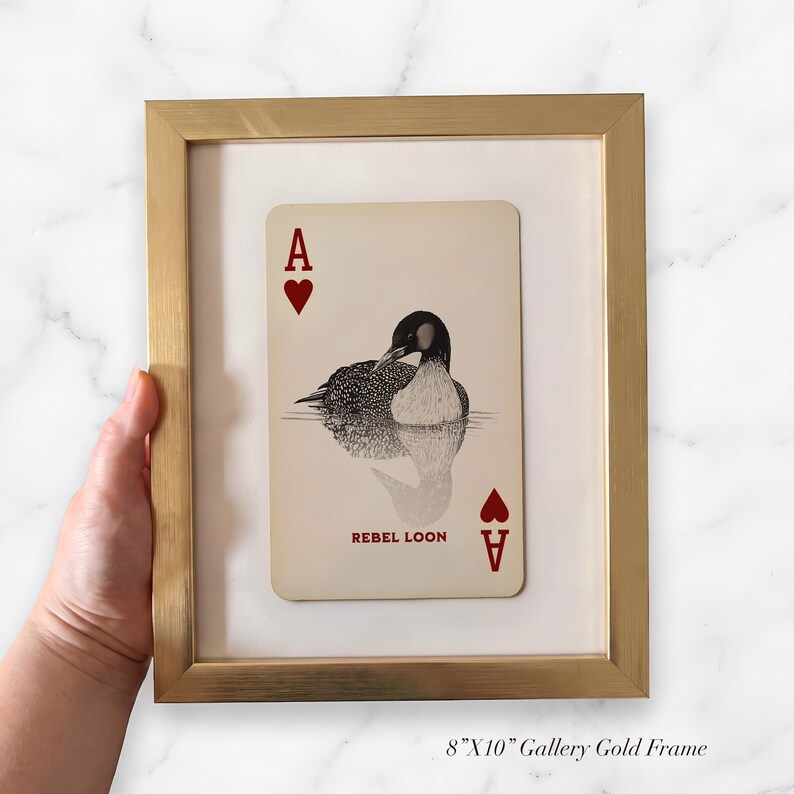 Puede incluir: Un marco de galer&iacute;a dorado de 20,32 cm por 25,4 cm que contiene una impresi&oacute;n de carta de juego. La carta presenta un dise&ntilde;o de as de corazones con una ilustraci&oacute;n en blanco y negro de un colimbo en el centro y el texto rojo REBEL LOON.